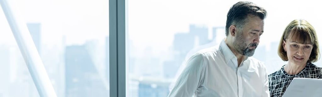 a man and woman in a high rise office, standing & looking at paperwork 