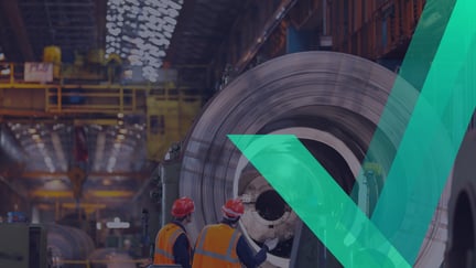 Two workers in orange safety vests and helmets inspect a large steel coil inside an industrial manufacturing facility, with machinery and overhead structures visible in the background.
